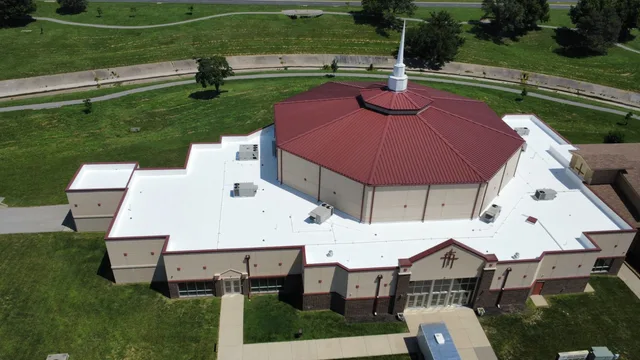 Aerial view of a church with a new red standing-seam metal roof over the sanctuary and white membrane roofing across the wings